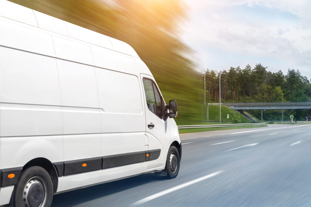 A white delivery van speeds down a highway with blurred trees in the background and an overpass in the distance. The sun casts a warm glow on the scene, highlighting the vehicle against the clear blue sky.